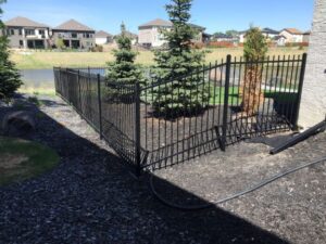 Genesis ornamental fence in Winnipeg installed on sloped residential yard with young trees and modern homes nearby